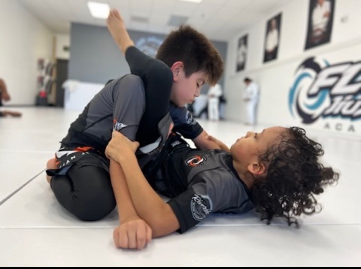 Dos niños trabajando la guardia en el tatami en una clase de jiu-jitsu No-Gi, con una clase de Gi entrenando atrás en Flow Academy Indio