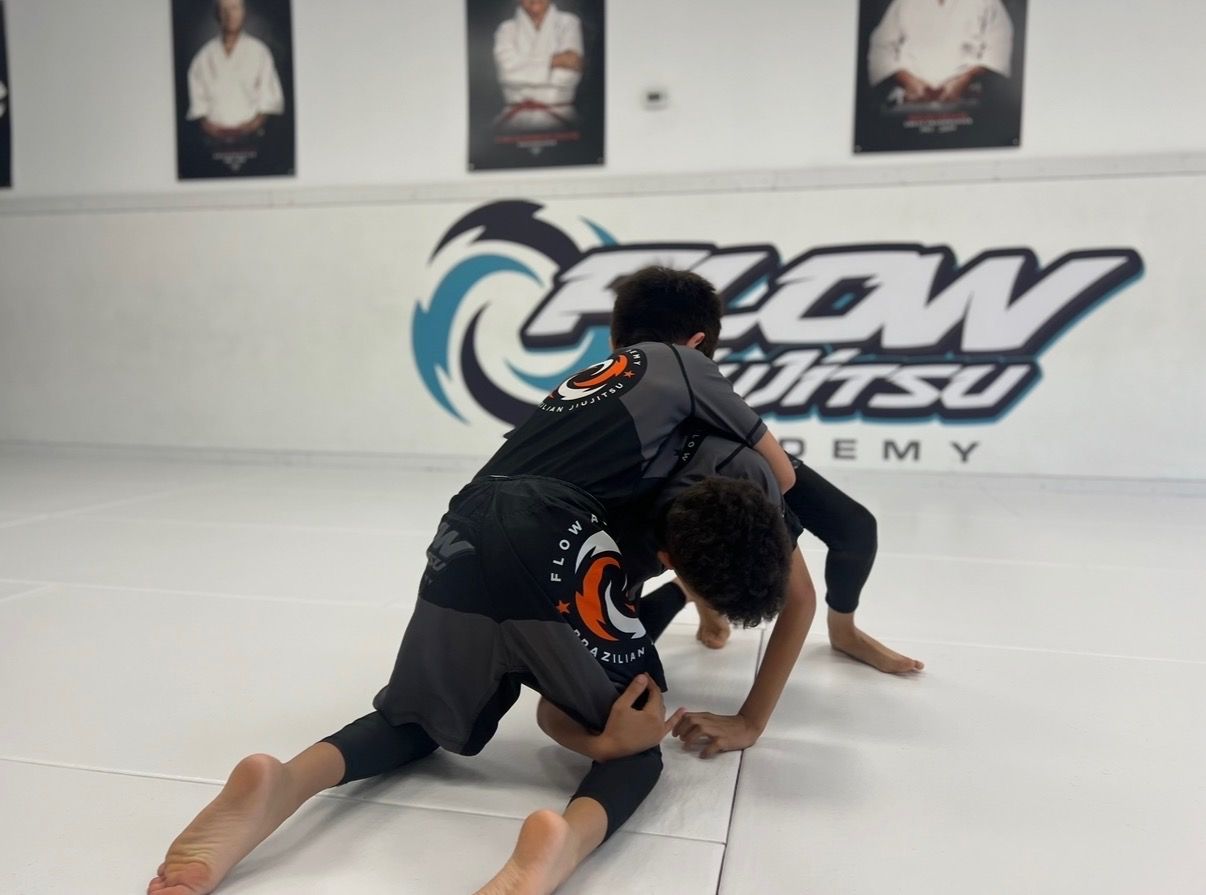 Dos niños con rash guard negro peleando por la posición en una clase de jiu-jitsu No-Gi frente al logo turquesa de Flow Jiu-Jitsu Academy en Indio