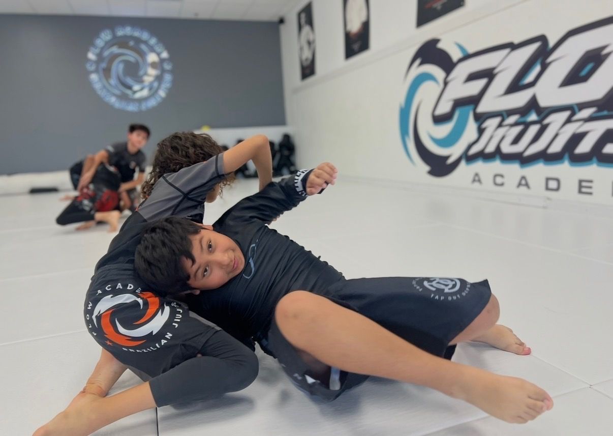 A smiling kid looks at the camera mid-drill during No-Gi jiu-jitsu while classmates train in the background at Flow Academy Indio