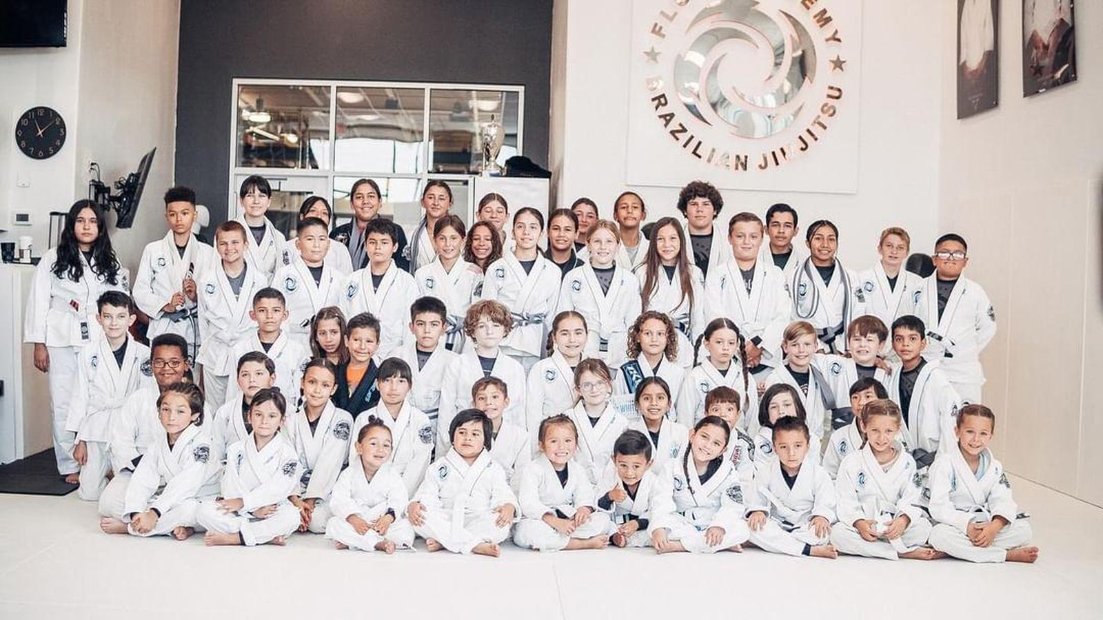 El equipo de niños y jóvenes de jiu-jitsu de Flow Academy Indio con gi blanco, sentados juntos en el tatami