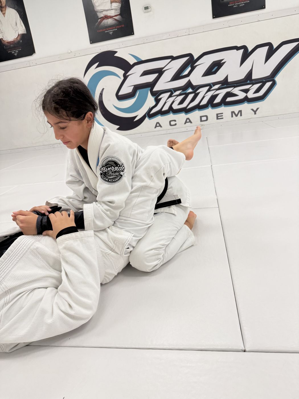 Una alumna juvenil con gi blanco, concentrada, terminando una llave de sumisión sobre su compañero en Flow Academy Indio