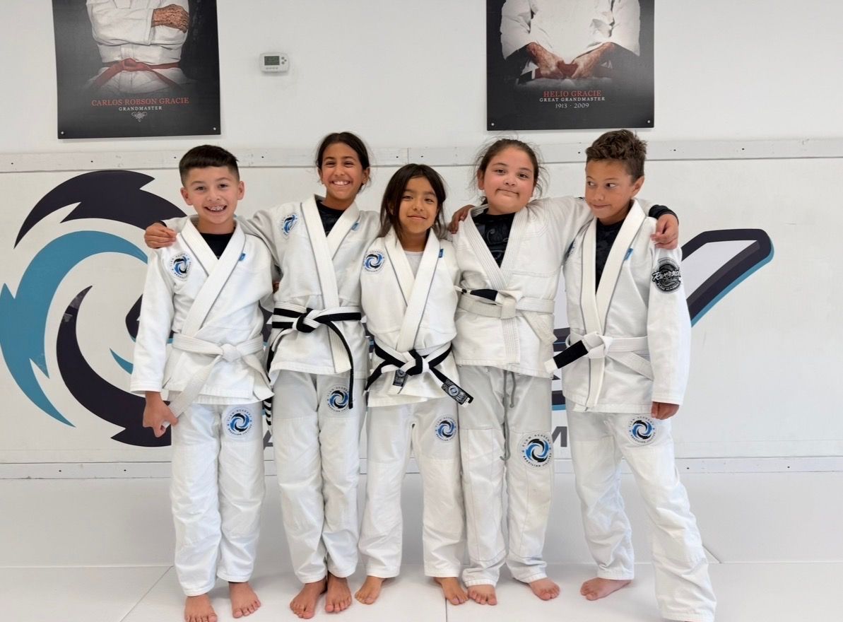 Cinco niños y jóvenes sonrientes con gi blanco de Flow Academy, de pie juntos en el tatami frente al logo turquesa de Flow en Indio